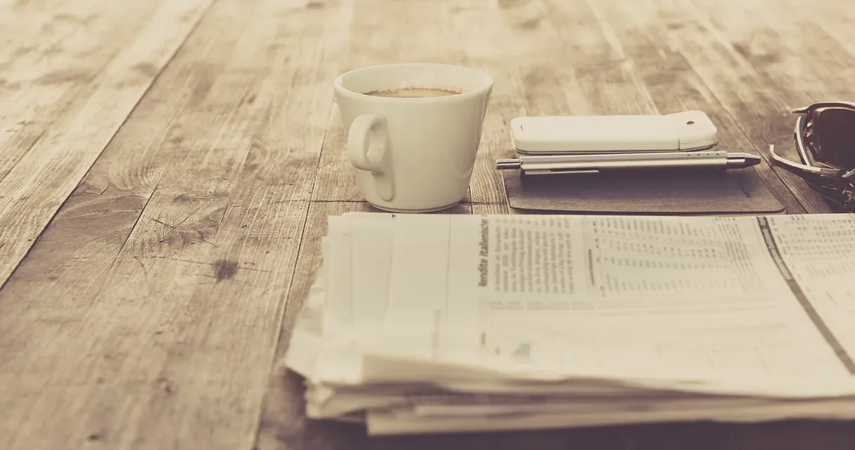 Xícara de café, celular e jornais empilhados sobre uma mesa de madeira