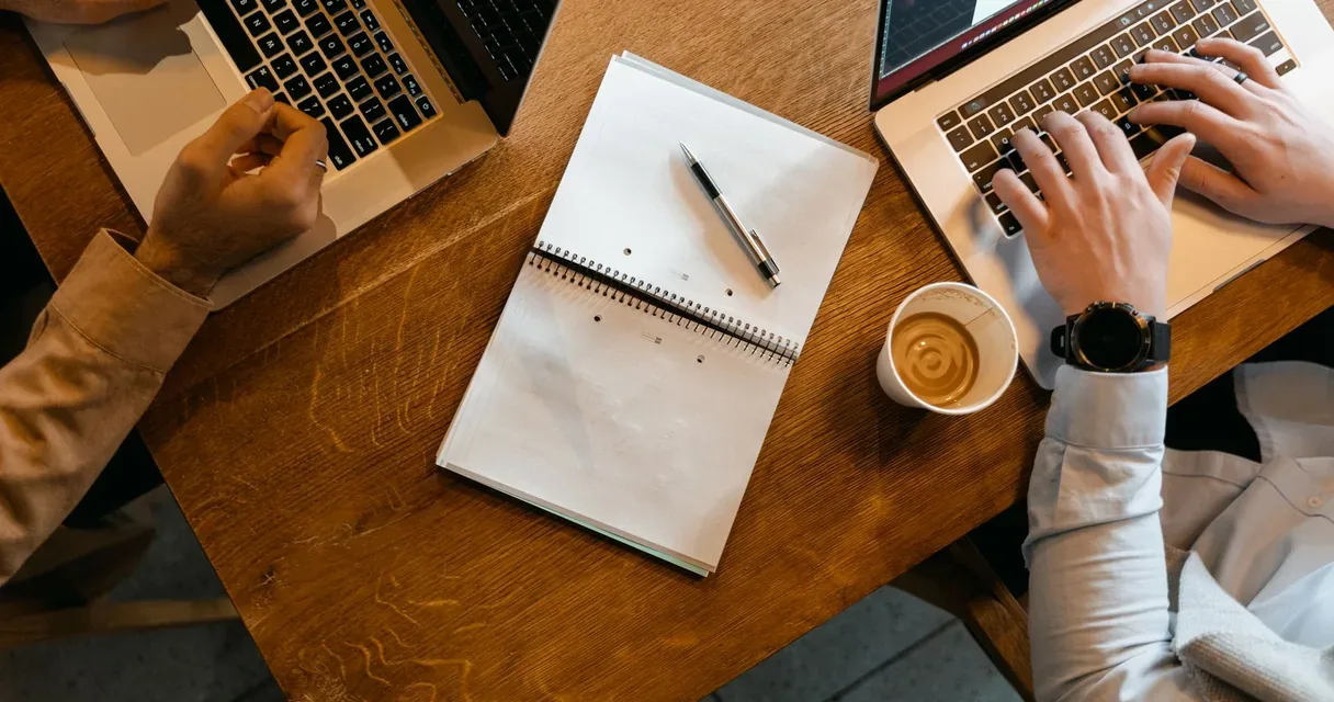 Duas pessoas digitando em notebooks lado a lado, com um caderno aberto entre eles