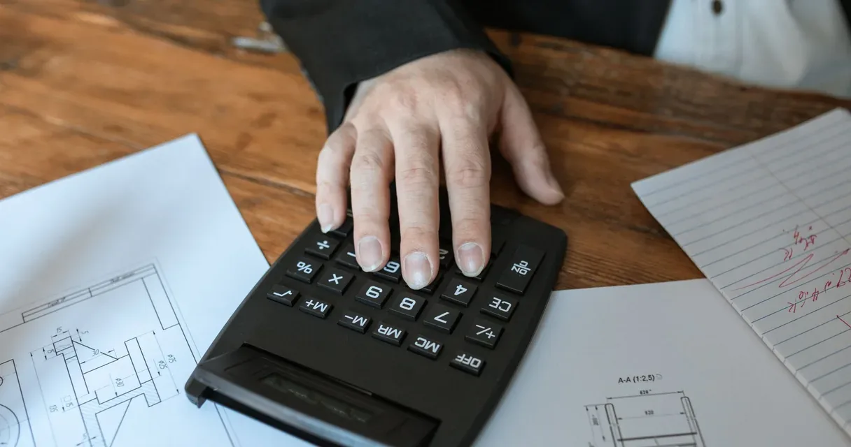 Mão sobre uma calculadora ao lado de papéis em uma mesa de madeira